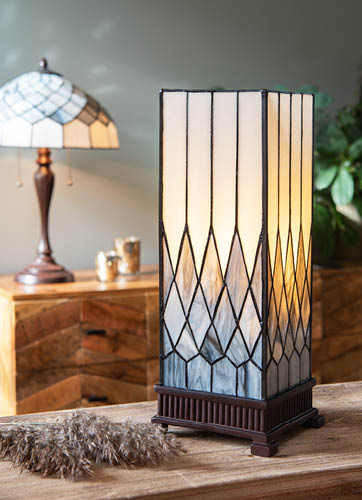 Een tafellamp te zien met een stijl van een van de Tiffany-lampen. Het heeft een rechthoekige, torenachtige vorm met glas-in-lood panelen die op een geometrische manier zijn ingedeeld. De kleuren van de panelen variëren van crème in het midden tot donkerblauw en zwart aan de zijkanten, waardoor een elegant kleurverloop ontstaat. De lamp staat op een houten tafel of kast en heeft een donker metalen frame aan de onderkant. Op de achtergrond is nog een ander soort Tiffany-lamp te zien met een meer traditioneel, koepelvormig ontwerp. De omgeving heeft een gezellige, warme sfeer met een huiselijke uitstraling, inclusief een houten kast en een plant in de hoek van de kamer.